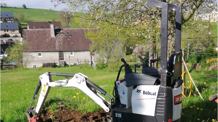 Auf dem Bild ist ein Minibagger Bobcat E10 mit einem Grabenräumlöffel abgebildet. Dieser zieht gerade die Oberfläche einer Wiese ab um diese zu begradigen.
