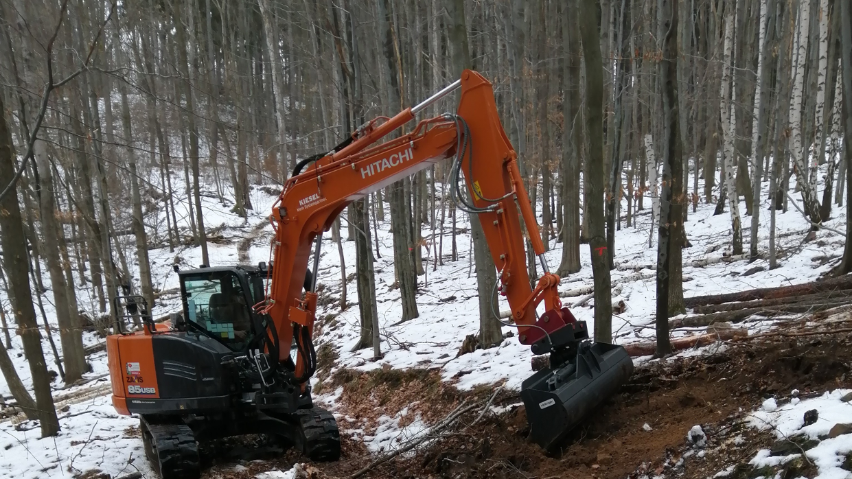 Auf diesem Bild ist der Bagger Hitachi ZX 85 mit dem Grabenräumlöffel zu sehen, welcher gerade dabei ist eine Rückegasse im Wald zu erschließen um diese befahren zu können.